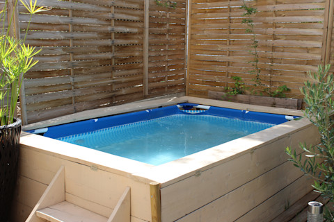A wooden framed pool with wood steps set up at the bottom of a garden in front of a wooden fence and surrounded by tropical plants.