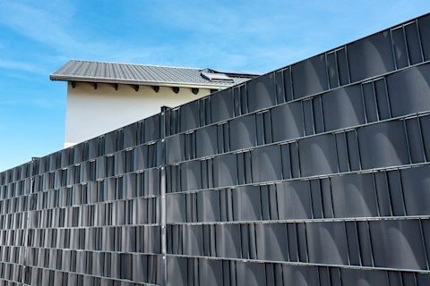 A large, metal garden fence with black privacy screen strips made of synthetic material bordering a home's garden. Part of the black roof and white exterior wall of the house appears above the fence, in front of a blue summer sky.