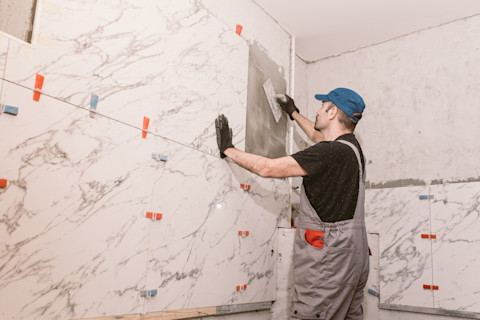 A construction worker in overalls and a blue cap applies adhesive to a tiled wall with a trowel. The bathroom has large white marble tiles, giving a clean, professional look.