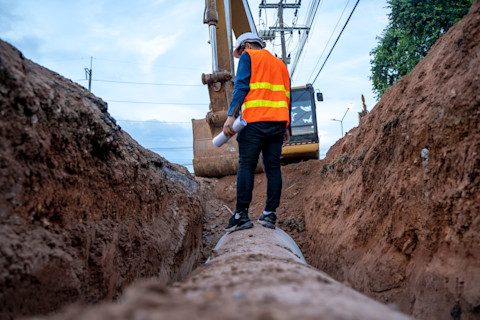 This image shows a groundworker wearing a high vis jacket and hard hat standing atop of a pipe in an excavated trench