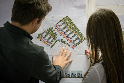 A man and a woman are attentively examining a colorful architectural site plan. Their fingers are placed on the paper, possibly pointing at specific details or areas of interest. The image shows the back of the man and over-the-shoulder view of the woman, both focused on the plan. The plan itself appears detailed with illustrations of housing units, landscaping, and layout annotations, indicating a residential development. The image captures a moment of professional collaboration and decision-making.