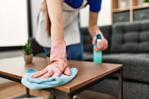 This image shows a cleaning professional wearing rubber gloves to clean a coffee table with a cloth. She has a cleaning spray in her left hand. 