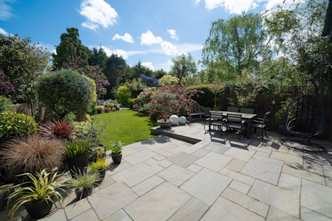 The image shows a lush green garden filled with plants, shrubs and trees and there is a paved patio. A wooden dining table sits on top of the patio, ready for alfresco meals.
