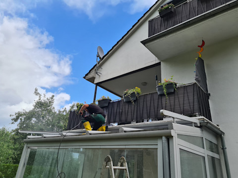 This image shows a tradesperson on top of a conservatory roof. He has accessed the roof via a ladder in the foreground and is using a tool to repair or clean the conservatory roof