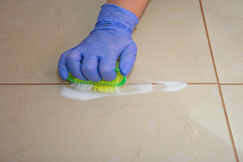 A person wearing blue gloves scrubs a tile floor with a green brush to remove grout, creating white soap suds, conveying cleanliness and meticulous care.