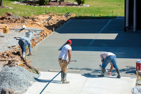 Three construction workers are smoothing wet concrete for a driveway on a sunny day. Gravel and equipment are visible nearby, with a grass lawn in the background.