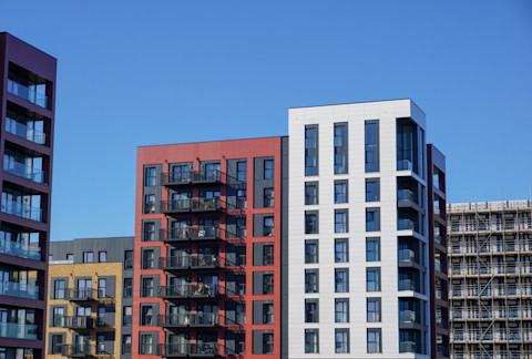 This image shows two modern high-rise residential buildings. The building on the left is covered with red cladding panels and the building on the right is covered with white cladding panels. They are set against a bright blue sky.