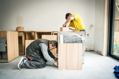Two workers are assembling wooden furniture in a bright, modern kitchen under renovation. The room has bare walls and a concrete floor, indicative of ongoing work. One worker, wearing a yellow shirt, is leaning over a cabinet, placing bricks on it, possibly to weigh it down or for spacing. The other, in a gray jacket and kneeling, is working on another section of the cabinetry. A wrapped countertop and various unpacked and half-assembled pieces suggest the kitchen is in the early stages of fitting.