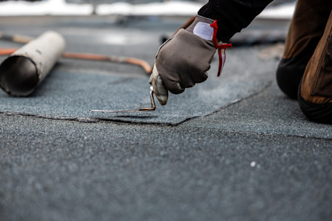 A worker in gloves uses a propane torch to lay roofing felt on a flat roof, with tools visible in the background.