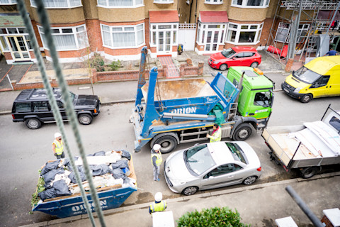 A busy street with many vehicles. A green and blue grab lorry drives by, and a skip full of rubbish sits nearby.