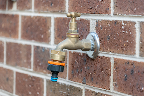 Close-up of an outdoor water tap mounted on a wall. The tap is shiny and new, with a hose attached.