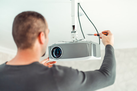 A man is in the process of setting up a projector in a living room, carefully connecting a cable to it.  The man is dressed in grey, and the living room has a clean, white backdrop, creating a bright and neutral environment that highlights the setup.