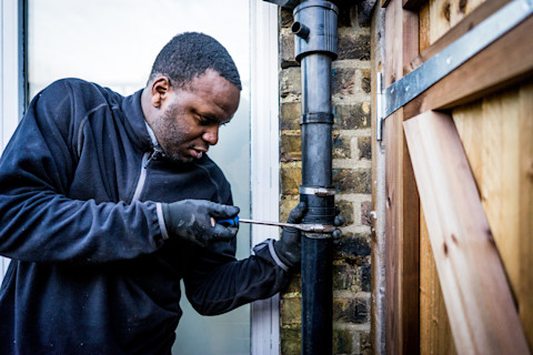This image shows a plumber wearing protective gloves using a tool to fix an external leaking pipe on the side of a house.