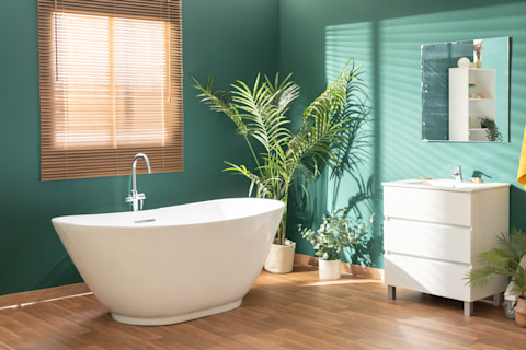This image shows a modern, brightly lit bathroom featuring green walls, a white freestanding bath, leafy plants and light wood accents.