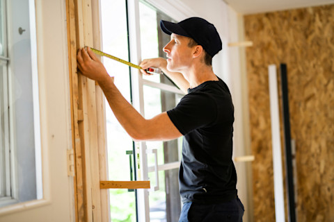 A young man in a black cap and t shirt measures the size of a patio door way, where one patio door is already installed, using a tape measure.