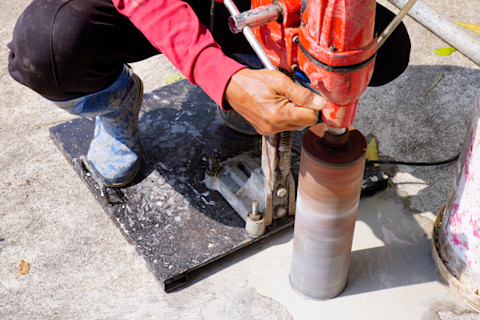 A construction worker performing core drilling on a concrete surface using a heavy-duty drill machine. The image highlights the drilling process, equipment, and setup, illustrating the typical work involved in core drilling projects, which affects overall costs