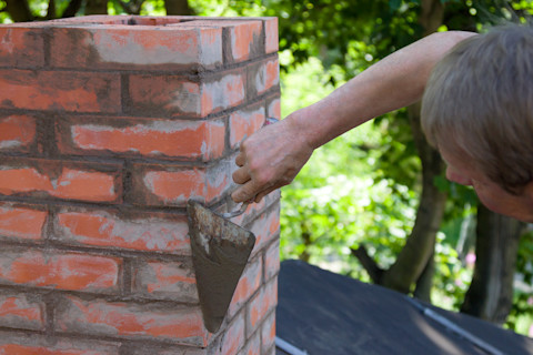 This image shows a chimney specialist or masonry expert using a trowel to repoint a red brick chimney stack. 