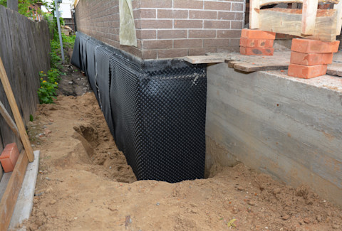 The image shows a construction site where a foundation wall is being waterproofed. The foundation is exposed, and a black dimpled waterproof membrane is applied along the exterior surface. The membrane helps prevent water infiltration into the building's foundation. The area around the foundation has been excavated to allow for the installation of the membrane. The exposed ground and soil are visible alongside the wall, and a temporary wooden structure is present on the right side, possibly for construction support. A wooden fence runs along the left side of the area.
