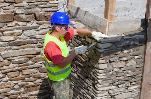 A stone wall being constructed, showing stone blocks being carefully placed and arranged, with masonry tools and partially completed sections visible. by a professional builder wearing a hi-vis vest and hard hat.