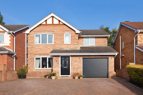 This image shows a modern brick house with two stories. The house has a dark-colored front door and a large window on the left side. On the right, there is a garage with a dark gray roller shutter door. The house is surrounded by neighboring properties, which have a similar design. The driveway is paved with light brown bricks, and potted plants are placed near the entrance, giving a welcoming touch. The sky is clear and blue, providing a bright, sunny backdrop for the scene.