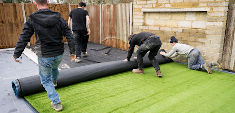 This image shows four landscapers working together to unroll artificial grass in a back garden. 