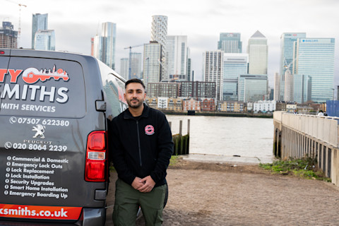 A locksmith standing confidently in front of his service van, which is adorned with the company branding 'City Locksmiths', displaying contact numbers and a list of services such as emergency lockouts and security upgrades.