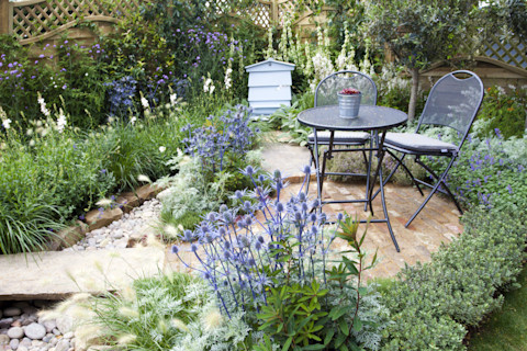 This image shows a beautiful landscaped patio with a small table and chairs and colourful flowers in varying hues of blue and purple, neatly trimmed shrubs and a curved stone pathway
