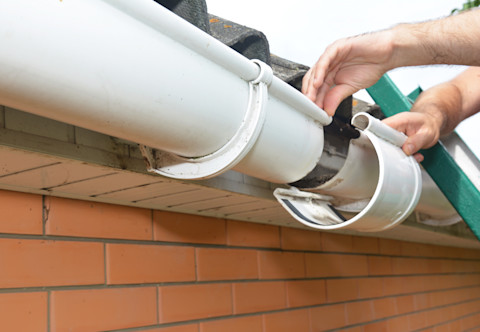 Hands repairing a detached section of white rain gutter on a brick house.