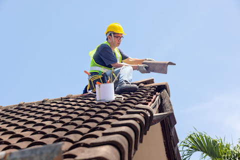 Worker repairing leaking ridge tiles on a tiled roof under a clear blue sky.

