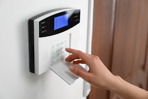 A female hand pressing numbered buttons on a white, digital burglar alarm security system pad, which is attached to the interior wall of a home beside the front door.