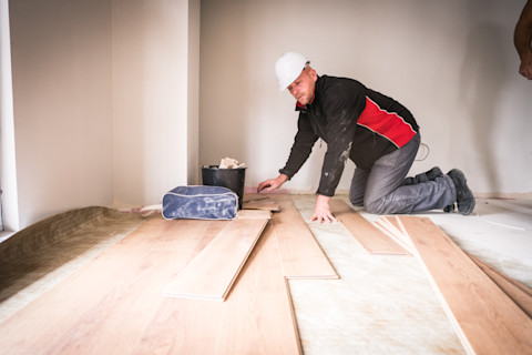 A construction worker, kneeling on a newly installed laminate floor, is in the process of laying down wooden planks. The worker wears a white hard hat, a black and red jacket, and gray trousers. In the foreground, a toolbox and a pile of wood planks can be seen. The room is spacious with white walls, creating a contrast with the warm tones of the wooden floor. Natural light from the window on the right illuminates the scene, casting soft shadows on the floor.