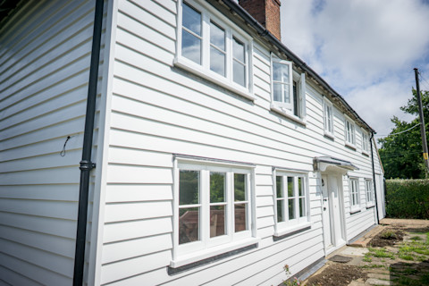 A two-story, traditional white clapboard house with black trim around white-framed windows, a white door, and a downspout on the left side, under a blue sky with clouds.