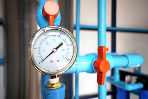 Close-up of a water pressure regulator installed on a plumbing system. The image shows the regulator with a gauge, connected to pipes. The background includes other plumbing components and fixtures.