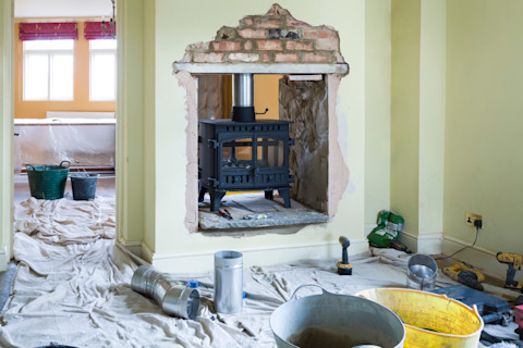 This image shows a woodburning stove being installed. There are dustsheets on the floor and part of the paint and plaster has been removed from the wall as part of the installation process.