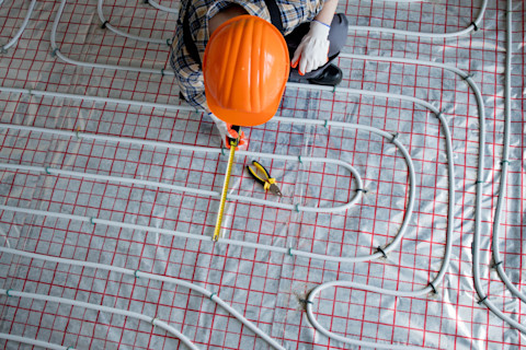 This image shows a tradesperson installing underfloor heating. They are wearing an orange hardhat and have an extended tape measure in hand. The heating components are visible as the flooring hasn't been installed yet.