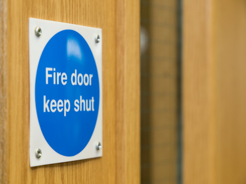 Close-up of a blue "Fire Door Keep Shut" sign mounted on a wooden door, highlighting essential fire safety precautions in a building.