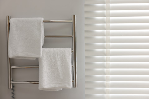 This image shows a chrome towel rail by a window covered by a closed Venetian blind. There are two white towels - one on the top rung and the second on the third rung. 
