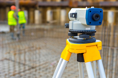 This image shows a close up of a Theodolite instrument on a construction site to establish boundaries of land. In the background two construction workers are wearing high-visibility vests.