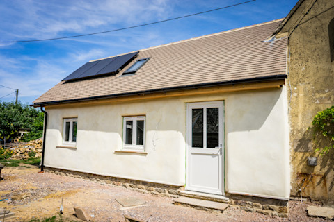 A newly renovated single-story house with a beige exterior, featuring a pitched roof with a row of solar panels. The house has two white-framed windows and a white door with a centered handle, all set against freshly plastered walls. The ground in front of the house appears to be under landscaping, with an uneven, dirt surface and construction materials scattered around. To the side, a portion of the old wall shows signs of wear and patchy plaster, contrasting with the renewed sections, suggesting ongoing exterior refurbishments.