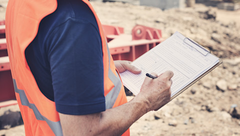 A man in a dark blue t shirt under an orange hi-vis jacket holding a clipboard and a pen, presumably making notes on the ongoing building work. He is standing in front of some orange building machinery and sandy ground.