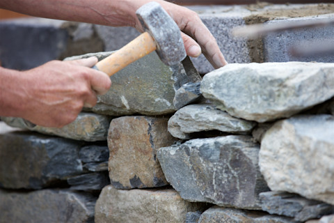 Close‑up of a stonemason’s hands using a hammer and chisel to shape and fit stones in a dry stone wall construction.