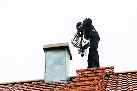 This image shows a chimney sweep standing on a residential rooftop with equipment, preparing to clean the chimney.