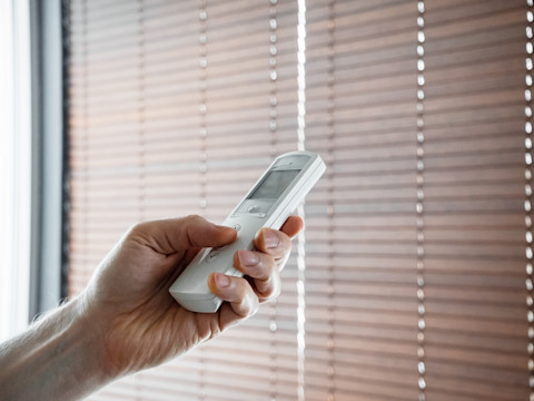 A hand is pointing a remote control at beige blinds, pressing the buttons. The blinds are closed, with sunlight gently filtering through the gaps.