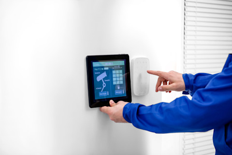 A hand in a blue jacket is pressing an iPad mounted on a white wall, with the device displaying the settings screen.
