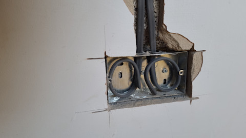 This image shows the initial stages of a double plug socket installation. A hole has been cut into the wall and the bracket has been placed into the space. There are two thick, black wires feeding into the area where the double plug socket will be installed. 