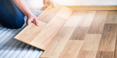 A person in blue overalls installing light wood laminate flooring on a grey underlay. 