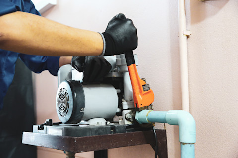 A man dressed in a blue jacket and black gloves is holding an orange tool while working on a pale blue water pump. He is adjusting something at the end of the pump.