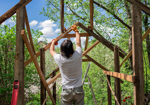 A man is midway through building a shed, with the skeleton of the roof in place. Using a tape measure, he measures one of the beams used to build the skeleton of the roof. Through the structure, leafy trees are visible against a bright blue sky.