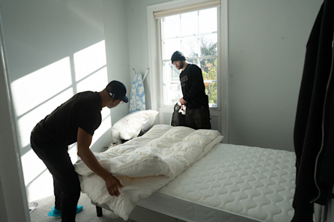 Two workers in a sunlit bedroom, one at the foot of the bed adjusting a white duvet on a bare mattress, and the other standing by the window, holding what appears to be a black bag. They are both wearing casual work attire and baseball caps, with the one by the bed bending over the task, and the one by the window looking down, possibly at a work-related device or checklist. The room has a clean, minimalist aesthetic, with a large window that lets in ample natural light, creating a serene atmosphere.