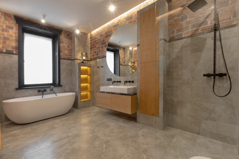 Loft interior of spacious wet room with bathtub near window and open walk in shower beside cabinet and sink with mirror against decorated built in shelves on gray marble
